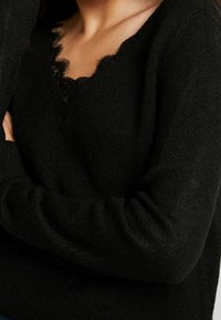 Czarny dzianinowy sweter z dekoltem w serek, ozdobiony koronkową lamówką. Materiał wydaje się miękki i lekko puszysty, z prążkowanymi mankietami.