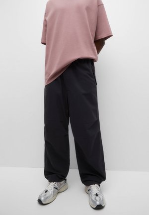 Personne portant un pantalon noir ample, une chemise mauve oversize à manches courtes et des baskets argentées, debout devant un fond blanc uni.