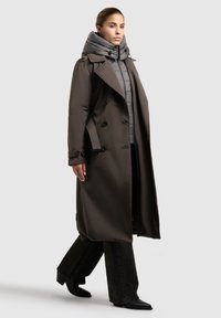 Kvinna som går, iklädd en lång mörkbrun trenchcoat över en grå pufferjacka, svarta vida byxor och svarta spetsiga stövlar.