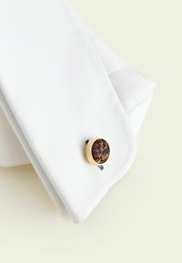 Närbild på manschett i texturerad vit skjorta med rund guldlänk med svart bakgrund och orange blommönster.