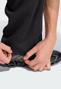 Ruce upravují černý elastický stahovací šňůrku na lemu černých kalhot nad černou sportovní obuví s vzduchovým odpružením.