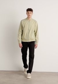 Hellgrüne Bomberjacke mit Reißverschluss, gerippten Bündchen und Saum, kombiniert mit schwarzen Hosen und beigen Schuhen. Glatte Textur, schlichtes Design.