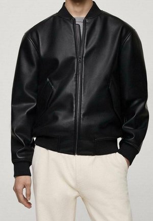 Mężczyzna ubrany w czarną skórzaną kurtkę bomberkę na zamek z prążkowanymi mankietami i pasem, w zestawie z kremowymi spodniami, z rękami w kieszeniach.