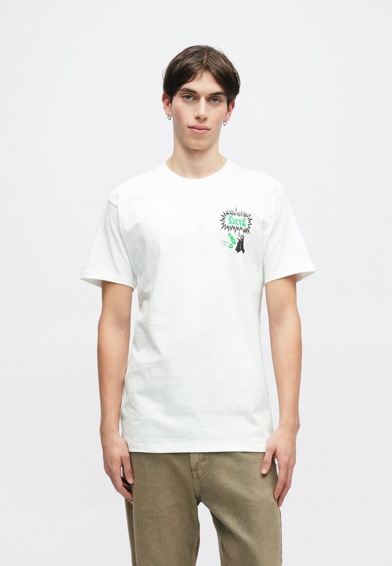 Λευκό βαμβακερό T-shirt με κοντά μανίκια, που διαθέτει πολύχρωμο γραφικό σχέδιο στο αριστερό στήθος, σε συνδυασμό με μπεζ παντελόνι.