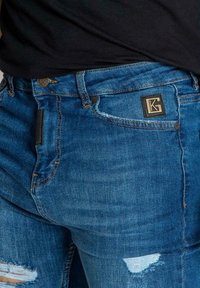 Sinise raitapuvut, joissa on kulunut muotoilu näkyvillä repeämillä, musta logopatchi ja kontrastiväriset ompelut yksityiskohdat.