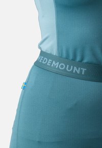 A teal athletic top featuring a soft texture, a wide waistband with "WE DEMOUNT" in light blue, and a small Swedish flag logo.