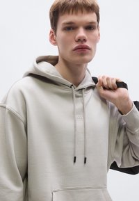 Helebeež värvi kapuutsiga dressipluus, millel on nööriga kapuuts, kängurutasku ja peen "BOSS" kaubamärgistus. Modell hoiab musta rihma õlal.