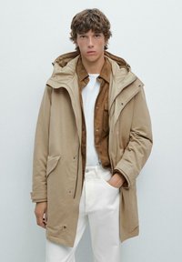 Beige vattentät parka med huood, med dragkedja och knäppning. Stylad över en brun skjorta och vita byxor. Slät tygtextur.