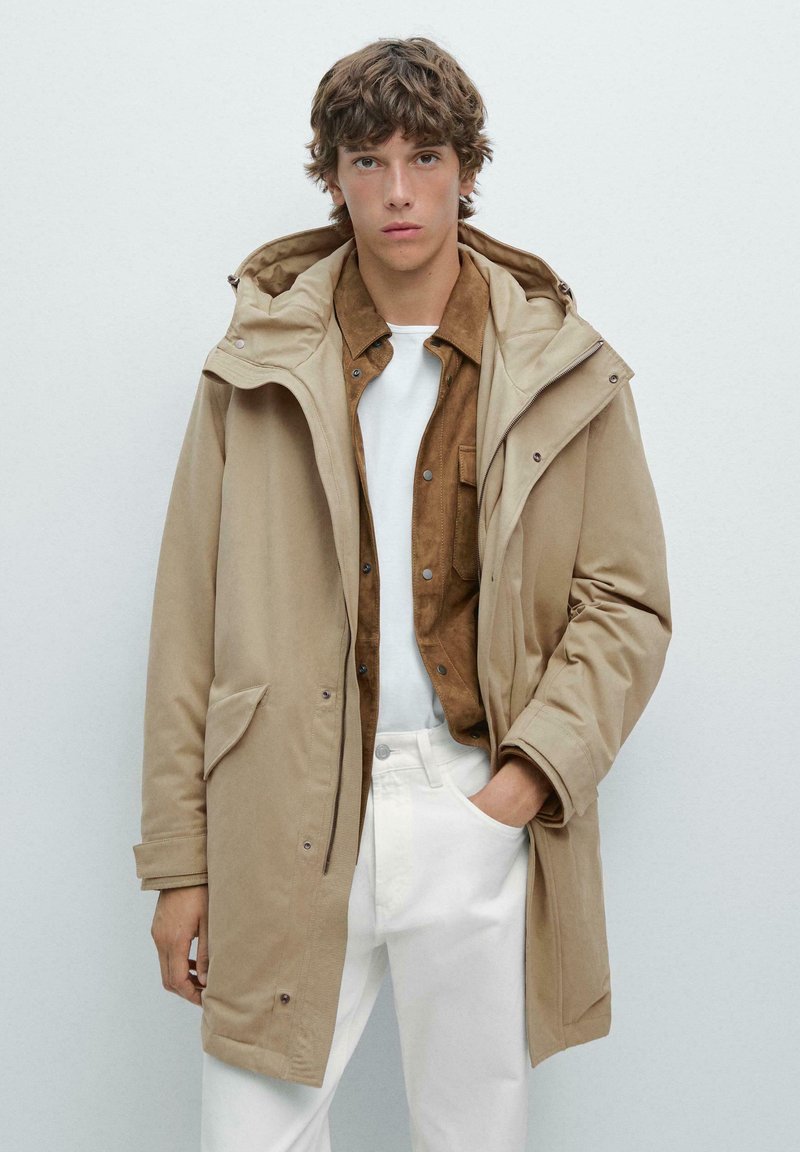 Beige vattentät parka med huood, med dragkedja och knäppning. Stylad över en brun skjorta och vita byxor. Slät tygtextur.