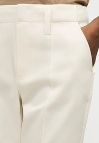 Κοντινό πλάνο ενός ατόμου που φοράει εκρού παντελόνι με το χέρι στην δεξιά τσέπη και μια μπεζ μπλούζα στερεωμένη μέσα στο παντελόνι.