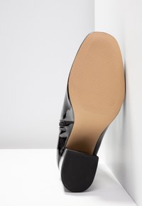 Black patent leather shoe with a rounded toe, featuring a textured rubber sole and a small, slightly elevated heel.
