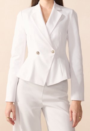 Femme portant un blazer blanc ajusté avec des boutons décoratifs et un pantalon blanc assorti, debout devant un fond neutre.