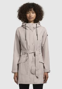 Parka imperméable beige clair avec taille cintrée, capuche et deux poches avant. Équipée de cordons de serrage à bascule et d'une finition lisse et texturée.