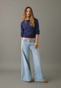 Mornarica pleten pulover, svetloplavi široki traper hlače visokega pasu, vidno šivanje in gladka tekstura. Obleko dopolnjujejo udobni čevlji.