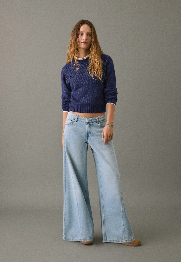 Mornarica pleten pulover, svetloplavi široki traper hlače visokega pasu, vidno šivanje in gladka tekstura. Obleko dopolnjujejo udobni čevlji.