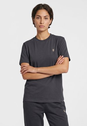 Persoon met kort donker haar die een donkergrijs T-shirt en bijpassende broek draagt met kleine lichte Chevron-logo's, staand met gekruiste armen.