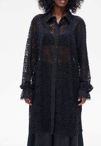 Czarna koronkowa bluzka z długimi rękawami i zapięciem na guziki z przodu. Posiada jednokolorowy czarny kołnierz i mankiety z falbankami. Przezroczysty kwiatowy wzór.