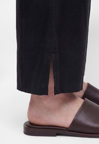 Lower leg wearing black pants with a small back slit above the ankle and brown slip-on flat shoes.