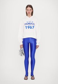 Hvit hettegenser med blå logo og tekst, kombinert med glinsende blå leggings, svarte sandaler med stropper og en sølvfarget håndveske.