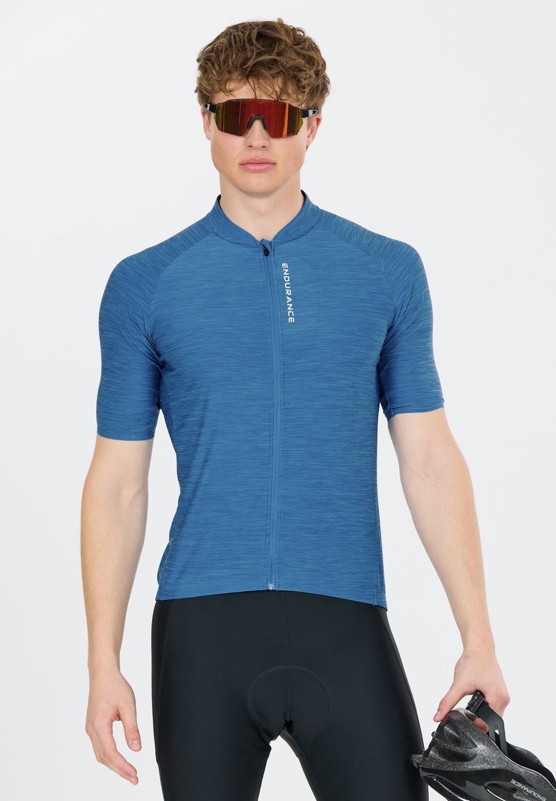 Homme portant un maillot de cyclisme bleu et un short rembourré noir, tenant un casque de vélo noir, portant des lunettes de sport teintées rouges, debout devant un fond blanc.