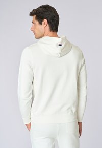 Felpa con cappuccio beige chiaro, maniche lunghe, polsini e orlo a coste. Presenta un piccolo logo tricolore sul retro del cappuccio. Materiale in cotone morbido.