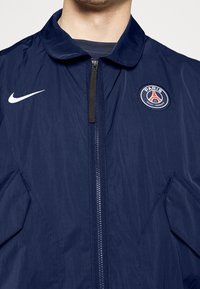 Laivastonsininen vetoketjullinen takki, joka on valmistettu kevyestä kankaasta. Takissa on Nike-logo ja pyöreä Paris Saint-Germain -patch rintataskussa.