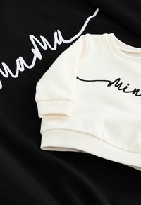 Czarna bluza z białym, kursywym napisem „mama” oraz kremowy top z pasującym napisem „mini” w czarnym, kursywnym stylu. Oba mają prążkowane mankiety i dekolty.