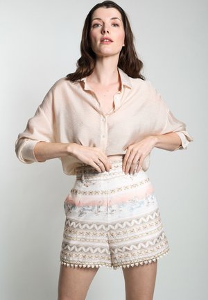 Lichtbeige overhemd met knopen en losse mouwen, gecombineerd met hoogsluitende shorts met een patroon in pastelkleuren en decoratieve accenten.