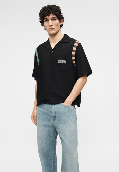 Νέος άνδρας με σγουρά μαλλιά που φοράει μαύρο πουκάμισο με κουμπιά και ριγέ μοτίβα στους ώμους, καθώς και ανοιχτόχρωμο μπλε τζιν, στέκεται με το ένα χέρι στην τσέπη.