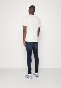Camiseta de algodón color beige claro, jeans de mezclilla oscuros de corte ajustado y zapatillas grises con detalles azules, vistos desde atrás.