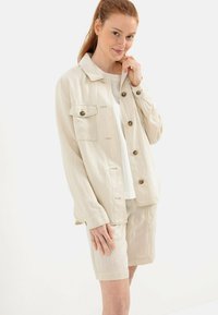 Lys beige jakke lavet af blødt stof med en knaplukning, der har to brystlommer og en afslappet pasform, parret med matchende shorts.