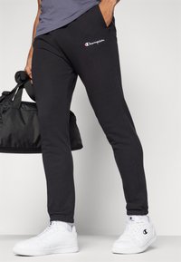 Pantaloni de jogging negri cu un design ajustat, fabricați dintr-un material moale. Prezintă un mic logo "Champion" roșu și alb pe coapsa stângă. Împreună cu adidași albi.