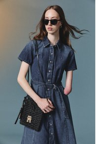 Denim shirt dress with short sleeves, button-down front, and waist belt. Paired with a black textured handbag and dark sunglasses.