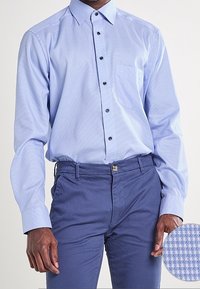 Uomo che indossa una camicia a maniche corte azzurra con un motivo, infilata in pantaloni blu scuro, mostrando un primo piano della texture del tessuto.