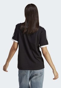 Černé bavlněné tričko s bílými akcenty na manžetách a trojicí pruhů na rukávech, zobrazené zezadu s volně střiženými džíny.