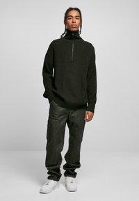 Pull décontracté noir en maille côtelée avec un col haut et une fermeture éclair à mi-hauteur, associé à un pantalon cargo gris foncé et des baskets blanches.