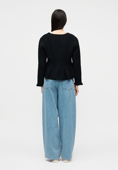 Γυναίκα με μακριά σκοτεινά μαλλιά, φορώντας μαύρο τοπ με μακριά μανίκια και βολάν στο τελείωμα, καθώς και ανοιχτόχρωμο τζιν παντελόνι με φαρδιές παντελόνες, που φαίνεται από πίσω.