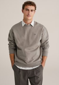 Svetlosivá mikina s žebrovanými manžetami a spodným lemom, s malým čiernym vyšívaným logom. Nosená cez bielu košeľu s golierom a kockované nohavice.