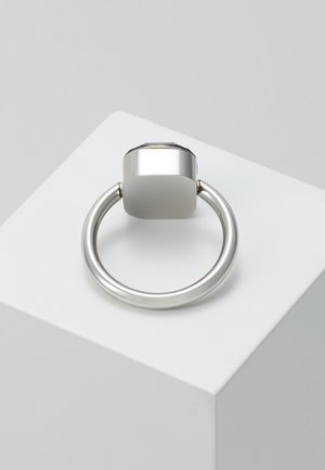 Srebrny metalowy uchwyt w formie pierścienia zamocowany do kwadratowej podstawy, umieszczony na białym sześcianie eksponującym na jasnoszarym tle.