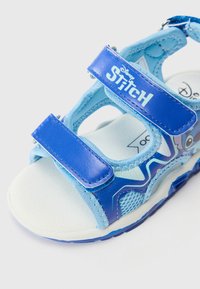 Siniset lasten sandaalit, joissa on verkko- ja synteettistä materiaalia, säädettävät hihnat ja teksturoitu valkoinen pohja. Mukana "Stitch" -brändäys.