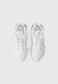 Sapatilhas Nike Shox brancas com uma parte superior texturizada, painéis em malha e detalhes em prata. Apresentam atacadores redondos e um colarinho acolchoado para conforto.