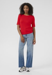 Rood, kort mouwen brei-top gecombineerd met lichtblauwe wijde jeans. Model draagt grijze sneakers; achtergrond is egaal wit.