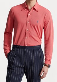 Korálová košile s dlouhým rukávem a knoflíkovým zapínáním, s klasickým límcem a vyšitým logem, v kombinaci s námořnickými pruhovanými kalhotami a hnědými hodinkami.