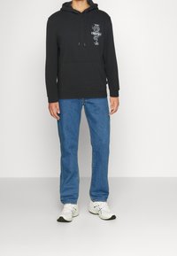 Hombre con sudadera negra con gráfico de dragón y texto, jeans azules y zapatillas blancas, de pie contra un fondo claro y liso.