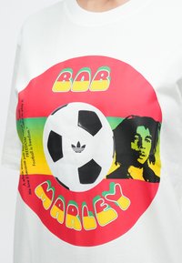 Λευκό πουκάμισο με κόκκινο, κίτρινο και πράσινο κύκλο που περιλαμβάνει μια ασπρόμαυρη ποδοσφαιρική μπάλα, το πορτρέτο του Bob Marley και το κείμενο «Bob Marley».