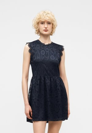 Mujer con cabello rubio rizado que lleva un vestido sin mangas de encaje negro, con corpiño ajustado y falda acampanada, sobre un fondo blanco liso.