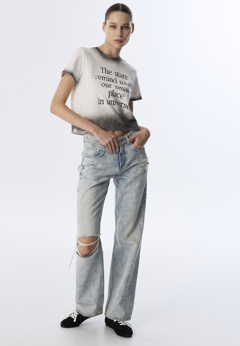 Geknipt shirt met een zwart-grijze verloop, bedrukt met tekst; lichtblauwe gescheurde denim jeans, rafelige zoom en zwarte sneakers met witte veters.