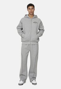 Felpa grigia con zip e tasche frontali, con cuciture nere a contrasto, abbinata a pantaloni della tuta grigi con linee di accento.