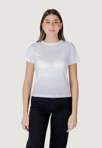 Witte T-shirt met korte mouwen, voorzien van een glanzend "GUESS" logo in lichtroze over de borst. Gepaard met zwarte broek.