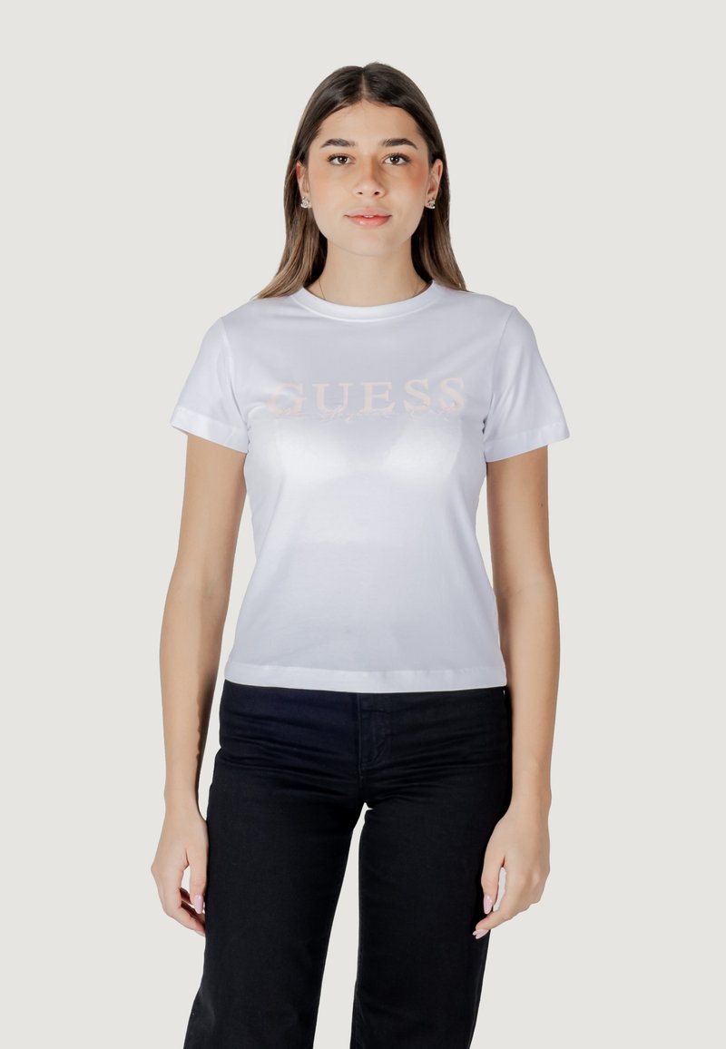 Witte T-shirt met korte mouwen, voorzien van een glanzend "GUESS" logo in lichtroze over de borst. Gepaard met zwarte broek.